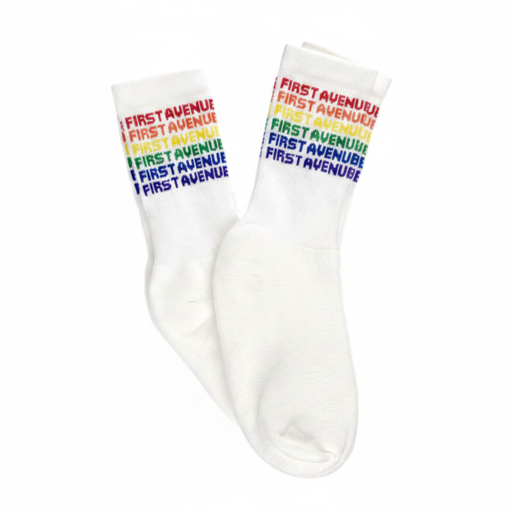 A pair of white crew socks with 'FIRST AVENUE' printed repeatedly in a rainbow color sequence on the cuff.